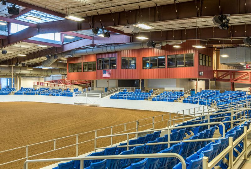 Indoor equestrian arena with sandy track, blue bleachers, and glass-encased VIP viewing area.