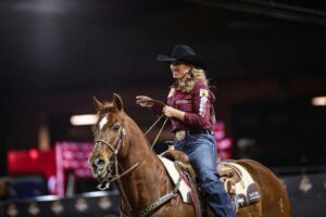 Confident cowgirl rides chestnut horse in indoor rodeo, showcasing Western skills.