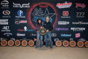 Rodeo champions celebrating with trophy amid sponsor logos at American rodeo event.